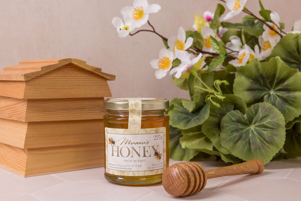 Jar of honey with a wooden honey dipper, bee hive-shaped container, and flowers on a light background