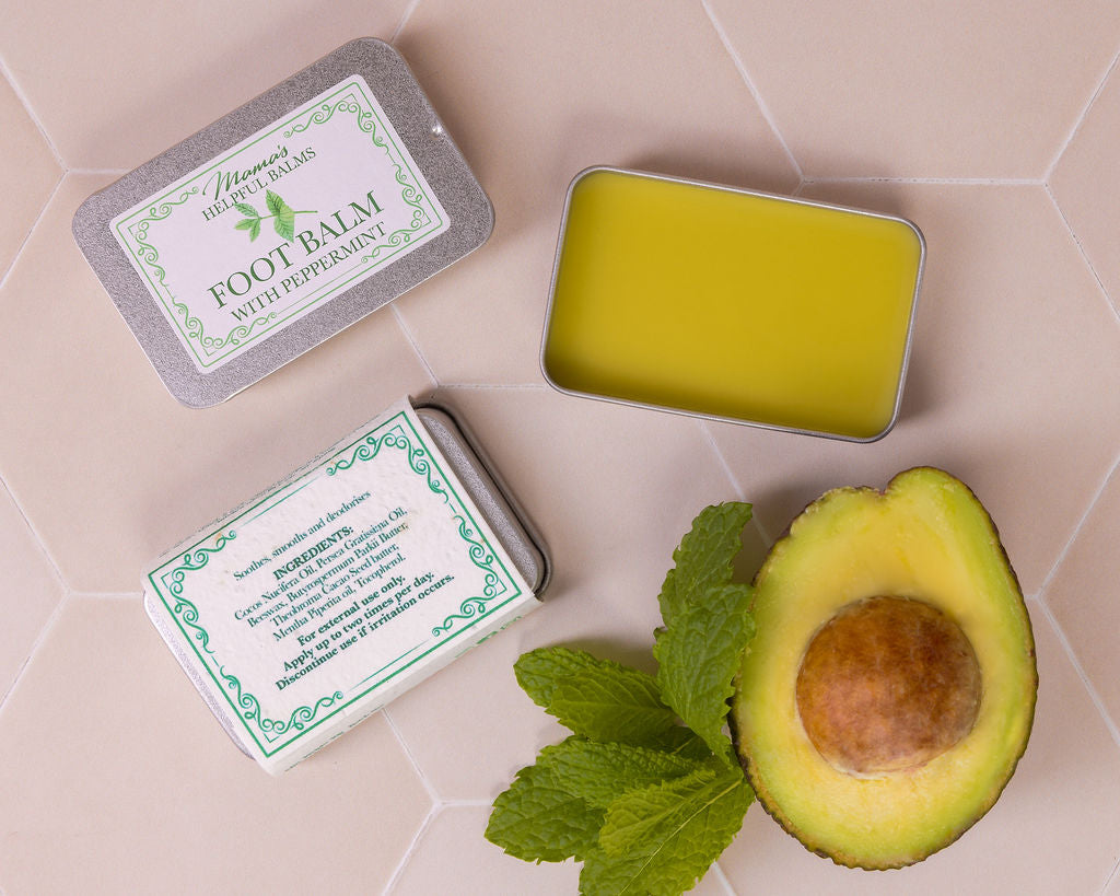 Mama's Helpful Foot balm in a tin with avocado and mint leaves on a tiled surface