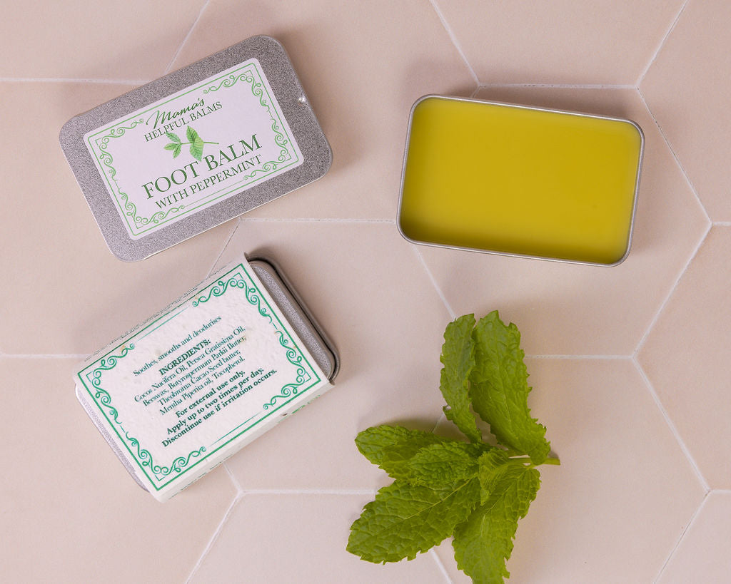 Mama's Helpful Foot balm with peppermint leaves in a tin on a tiled surface